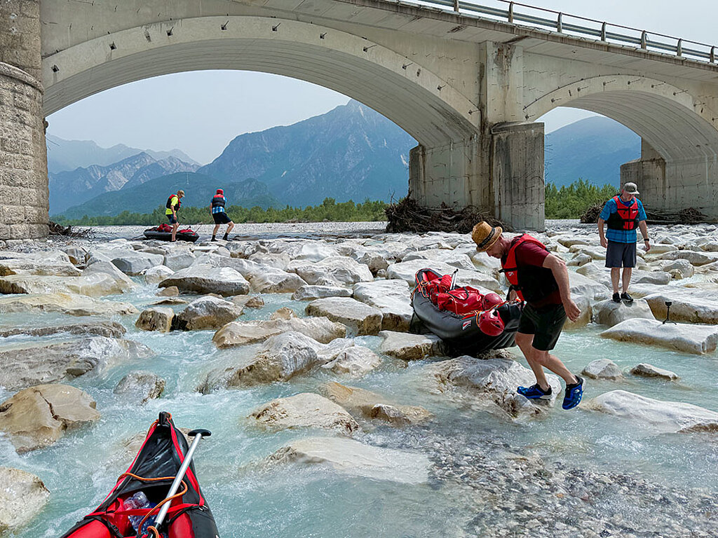 eine Gruppe Männer zieht ihre Schlauchboote über die Steinen von einem seichten Fluss